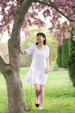 İlkbaharda çekilmiş canlı pembe çiçeklerle kaplı bir ağacın altında gülümseyen beyaz elbiseli bir kızın portresi..