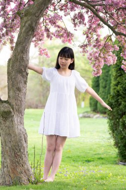 İlkbaharda çekilmiş canlı pembe çiçeklerle kaplı bir ağacın altında gülümseyen beyaz elbiseli bir kızın portresi..