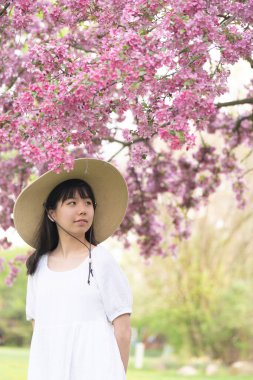 İlkbaharda çekilmiş canlı pembe çiçeklerle kaplı bir ağacın altında gülümseyen beyaz elbiseli bir kızın portresi..