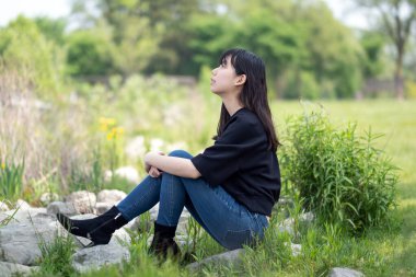 Genç Asyalı genç kız yeşil bitkiler ve kır çiçekleriyle çevrili çimenli bir alanda yerde oturuyor, siyah bir bluz ve kot pantolon giyiyor..