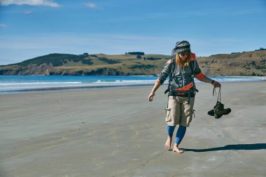 Sırt çantalı bir kadın Yeni Zelanda 'daki kumlu plaja geliyor ve okyanus kenarındaki botlarını çıkarıyor.
