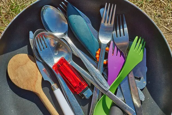 A pile of kitchen utensils in a pan outdoors in a camping. Forks, spoons, knives, some plastic, some metal, clean washed