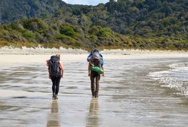Sırt çantalı bir adam ve kadın Yeni Zelanda, Rakiura Stewart Adası 'ndaki kumlu plaja doğru yürüyorlar.