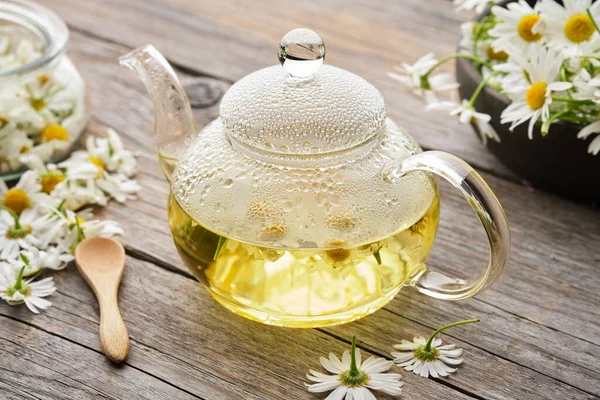 Glass tea kettle of healthy chamomile herbal tea. Chamomile daisy flowers on wooden table. Alternative herbal medicine.