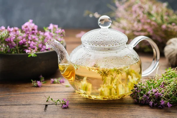 Glass tea kettle of healthy herbal thyme tea, medicinal herbs, bunches of healing thyme and heather flowers on table.  Alternative herbal medicine.