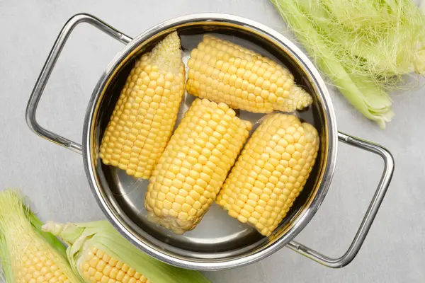 Cooking pot of yellow sweet corn cobs prepared for boiling, raw corncobs for cooking on kitchen table, top view.