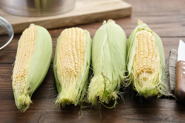 Raw sweet corn cobs for cooking and kitchen knife on kitchen table, cooking pot on background.