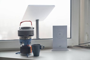 KYIV, UKRAINE - JANUARY 24, 2023: Close-up of Starlink antenna and wi-fi router on windowsill with portable burner stove with kettle and cup of tea beside.