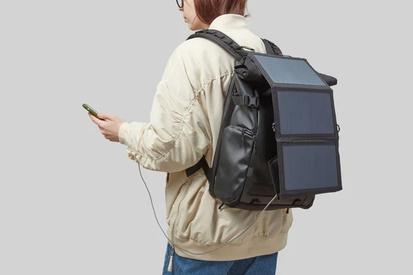 彼女の手にスマートフォンに取り付けられた柔軟なソーラーパネルを持つバックパックを運ぶ若い女性は ライトグレーの背景に隔離されました 旅行とグリーンエネルギーの概念 — ストック写真