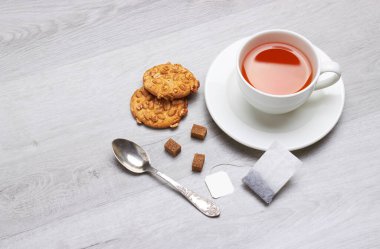 Bir fincan çay ve çay poşeti, kaşık, kurabiye, şeker, hafif ahşap arka planda.