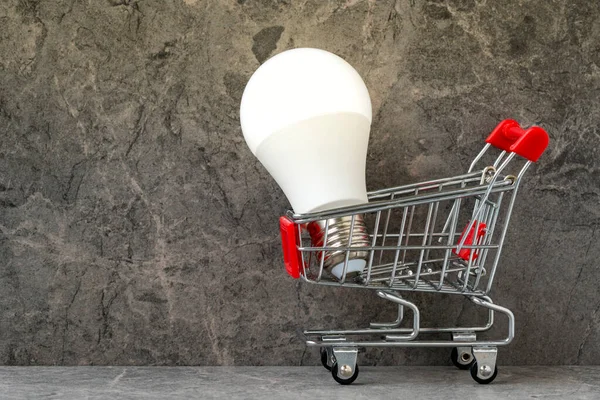 灰色の花崗岩の背景に明るい光る電球を持つショッピングカート 省エネ エネルギー価格の概念 — ストック写真
