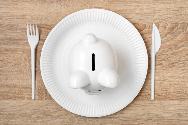 Piggy bank on the disposable plate with fork and knife, savings consumer concept. Spending savings, food expenses.