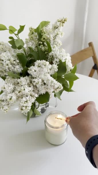 女人们用手在透明的玻璃瓶里点燃蜡烛 白色的紫丁香花束在白桌上 垂直拍摄的蜡烛视频模仿 真正的白色房间内部特写镜头 — 图库视频影像