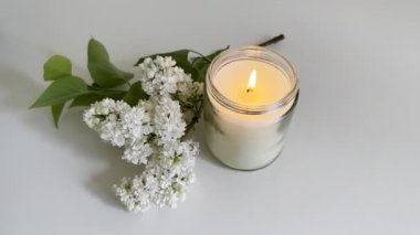 Temiz cam kavanozda yanan mum ve beyaz masada beyaz leylak buketi, gerçek beyaz oda içi yakın çekim