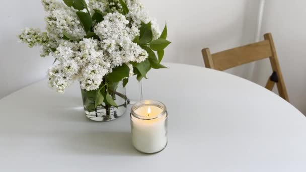 在透明的玻璃瓶中燃着蜡烛 白桌上点缀着白丁香花 制作了烛光视频模型 拍摄了真正的白色房间内部特写 — 图库视频影像