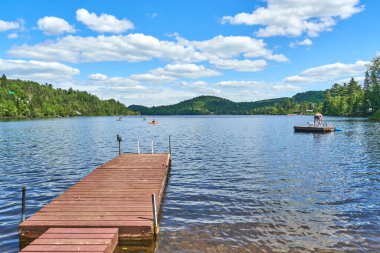 Sakin göl, kabarık bulutlarla benekli parlak mavi bir gökyüzünü yansıtır. Güneş ışınları, sakin suya uzanan uzun tahta bir iskelede dans eder. Gölde görünür kayıkçılar ve kayıkçılar var.