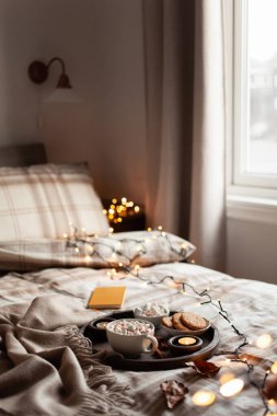 Sıcak sıcak yatak odası içi tepside bir fincan sıcak çikolata, mumlar noel ışıkları