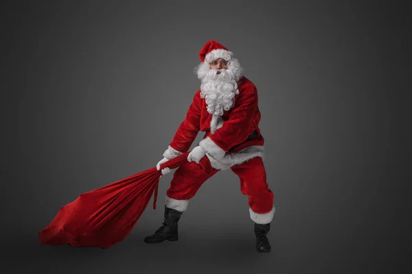 Shot of bearded santa with big bag stuffed with gifts against grey background.