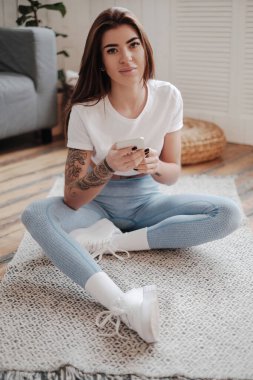 Cazibe portresi ve elleri dövmeli genç bir kız rahat bir oturma odasında telefonla yerde poz veriyor..