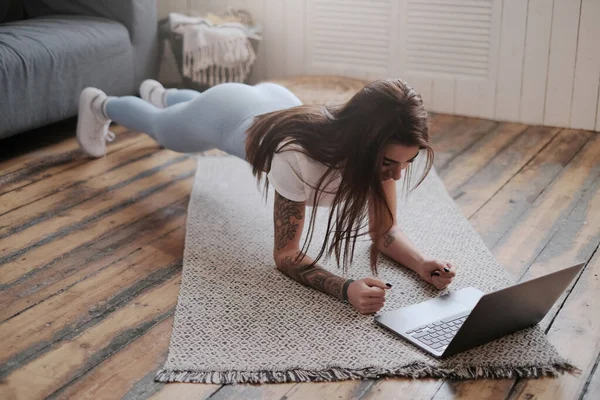 現代のラップトップを持つ入れ墨の女の子の健康的なライフスタイルは アパートの床に物理的な演習を行います — ストック写真