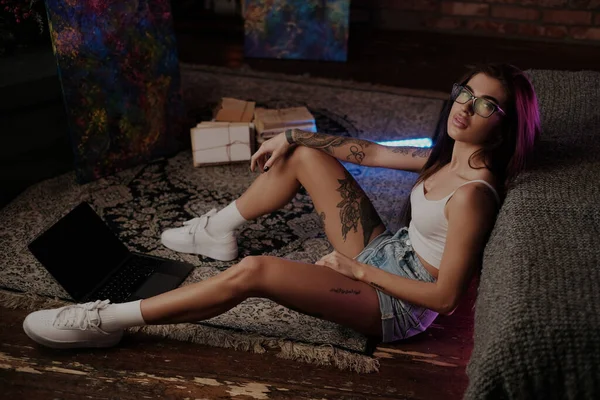 一个戴着眼镜 膝上型电脑在霓虹灯周围地板上的迷人姑娘的大气画像 — 图库照片