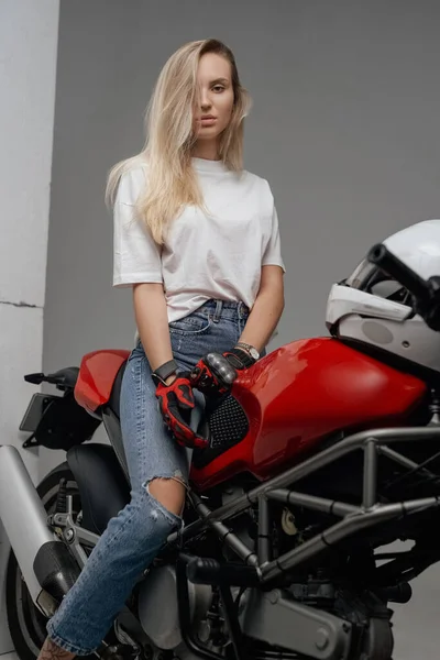 金发女郎穿着衬衫和牛仔裤骑红色摩托车的工作室照片 — 图库照片