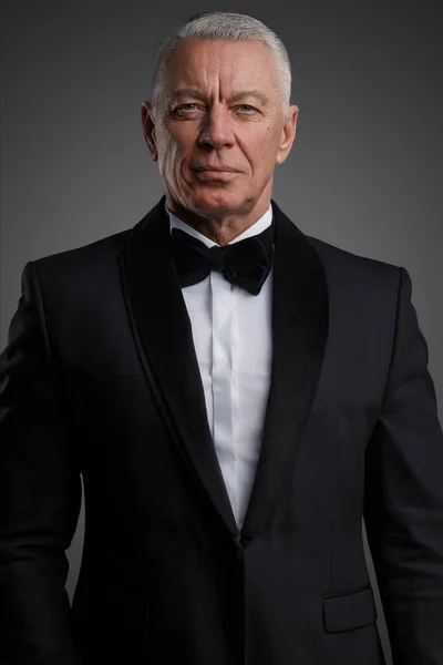 Distinguished older gentleman exudes confidence in this photo, dressed in a sharp black suit and bow tie. The grey background highlights his striking grey hair and determined pose