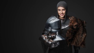 Beautiful female knight wearing medieval armor, smiling widely and confidently posing with a fur draped over one shoulder and a helmet in hand, on a gray background clipart
