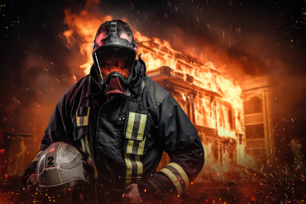 Мужественный пожарный в защитном снаряжении и кислородной маске стоит в окружении пламени и искр перед горящим зданием