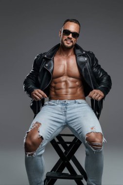 Kaslı gömleksiz yakışıklı erkek model geniş bir gülümseme sergiliyor. Siyah güneş gözlüğü, yırtık kot pantolon ve siyah deri ceket giyiyor. Stüdyo sandalyesinde gri arka planda oturuyor.