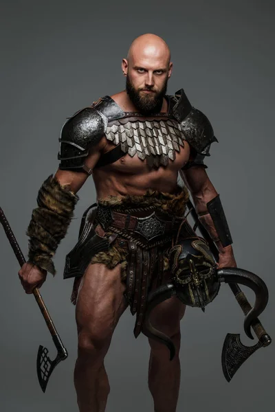 A bald bearded viking in lightweight fur-lined armor holding a two axes stands against a gray background
