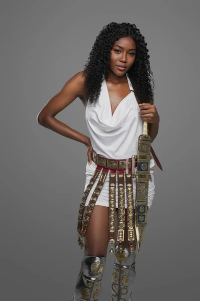 优雅的非洲裔美国女人 身披希腊华丽的白色腰带 头戴灰色背景 自信地拿着一把角斗士剑 — 图库照片