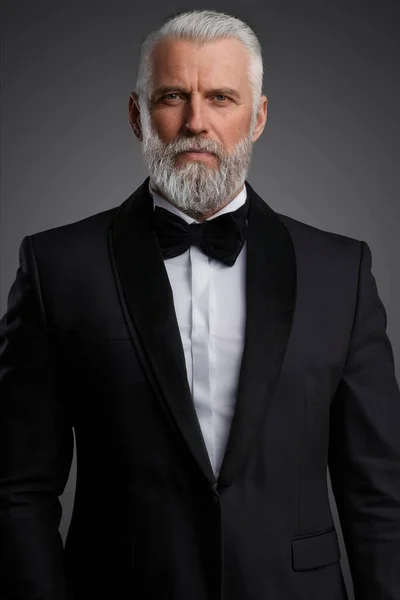 英俊而老练的绅士穿着雅致的黑色西服 打着领带 在拍照时 在灰色背景下传达着一位和蔼可亲的密探或间谍的形象 — 图库照片
