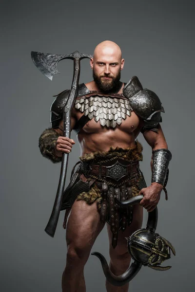 A bald bearded viking in lightweight fur-lined armor holding a large two-handed axe stands against a gray background