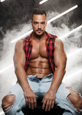 Kaslı, çıplak gövdeli, yırtık kotlu ve açık ekose gömlekli karizmatik erkek model rahat bir poz içinde, neon ışıklı dumanla çevrili.