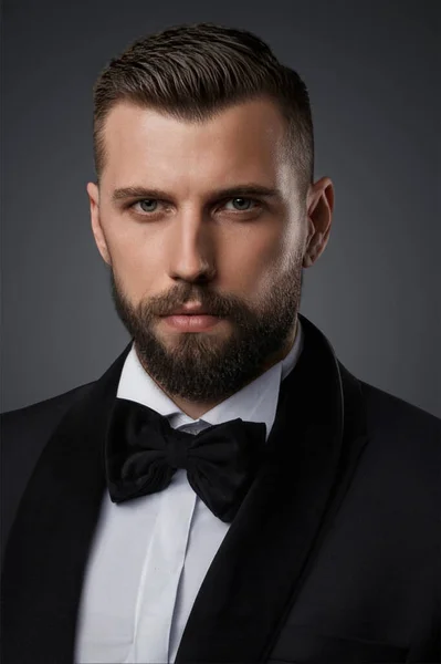 stock image Close-up portrait of a stylish and attractive man with a well-groomed beard, dressed in a high-end black suit and bow tie, posing confidently on a gray background