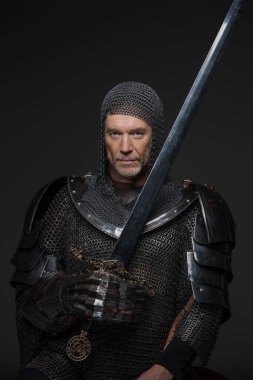 Gri sakallı, ağır zırhlı, gri bir arka planda kılıç tutan olgun bir kralın kendinden emin portresi.