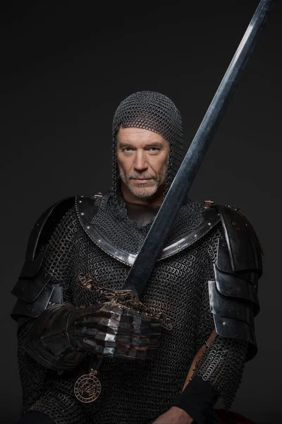 Confident portrait of a mature king with a gray beard, wearing heavy armor, holding a sword against a gray background
