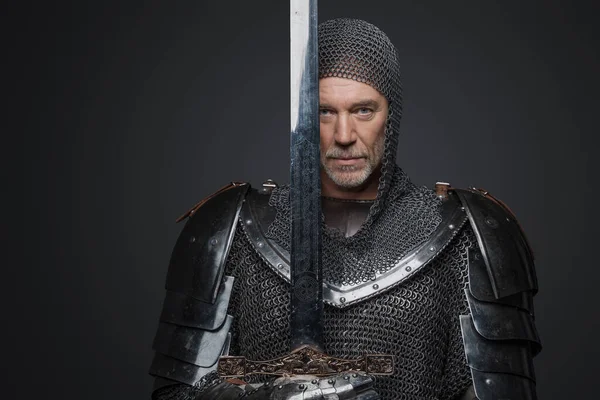 一个头戴灰色胡须 身穿厚重盔甲 拿着一把灰色背景的剑的成熟国王的自信画像 — 图库照片