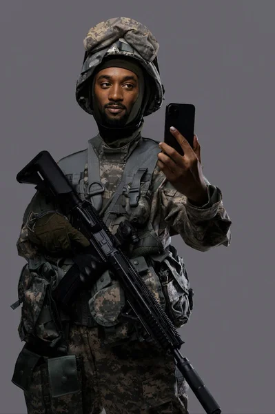 Natoの制服とヘルメットの暗い肌の兵士は 軍事要員の多様性と技術スキルを示す プレーングレーの背景に自撮りやビデオチャットのための彼のスマートフォンを保持しています — ストック写真