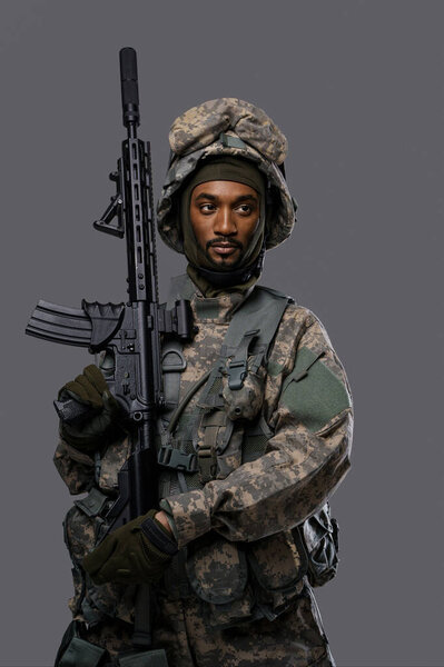 Dark skinned soldier in NATO uniform and helmet poses with a serious expression on a plain grey background