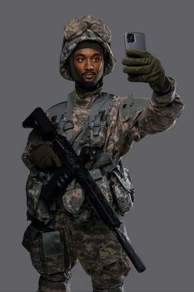 Natoの制服とヘルメットの暗い肌の兵士は 軍事要員の多様性と技術スキルを示す プレーングレーの背景に自撮りやビデオチャットのための彼のスマートフォンを保持しています — ストック写真