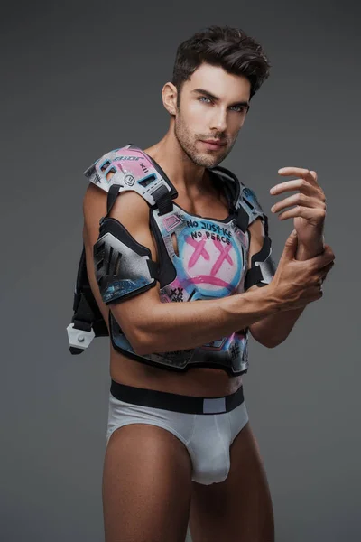 A portrait of a brunette fashion model posing in lingerie and wearing a street art inspired, grunge-style hockey protective gear, set against a gray background