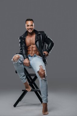 Kaslı gömleksiz yakışıklı erkek model yırtık kot pantolon ve siyah deri ceketle poz veriyor. Stüdyo sandalyesinde otururken gri arka planda seksilik ve güven yayıyor.