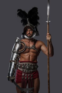 Güçlü ve zarif gladyatör, hafif zırhı ve tüylü kaskıyla tarafsız bir arkaplanda mızrak taşıyor.