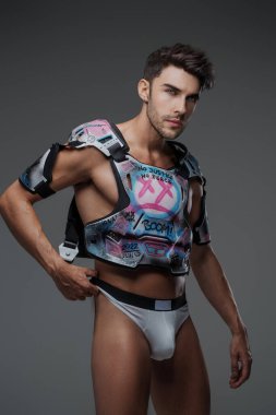 Koyu renk saçlı ve anlamlı özellikleri olan bir erkek manken, tarafsız gri bir arka plana karşı hokey esinlenilmiş koruyucu giysiler ve baştan çıkarıcı iç çamaşırlarıyla poz verir.