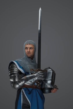 Gururlu şövalye elinde kılıcıyla miğferini, mavi paltosunu, zincirli paltosunu ve ortaçağ zırhını gri bir arka planda tutuyor.