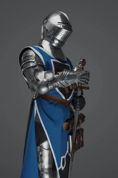 中世の兵士は青のコートと鎧に身を包み 灰色の背景を背景に静止した像のようなポーズの剣を持っていた — ストック写真