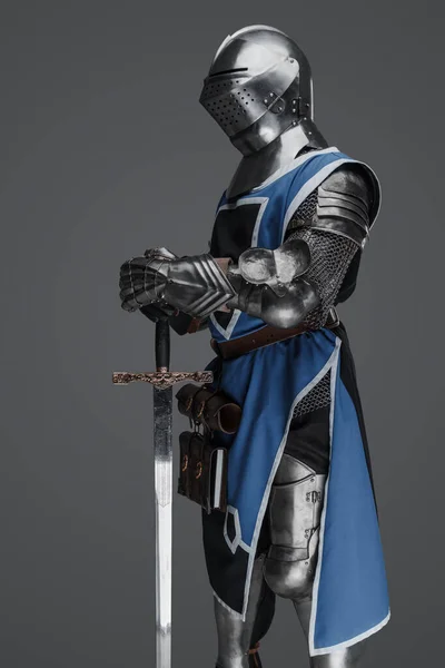 中世の兵士は青のコートと鎧に身を包み 灰色の背景を背景に静止した像のようなポーズの剣を持っていた — ストック写真
