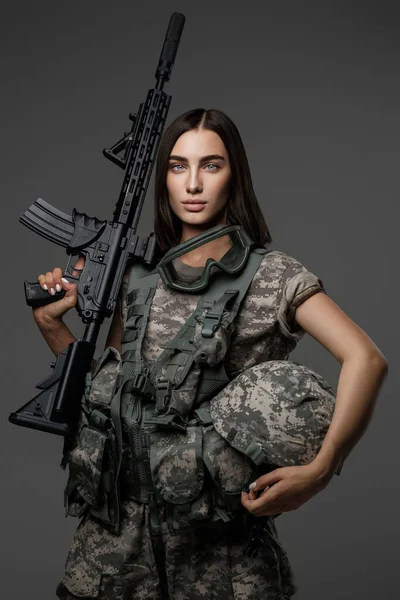 見事な青い目を持つ美しい女性兵士は 灰色の背景に撮影されたスタジオの肖像画のための自動ライフルを保持する彼女の軍服に立っています — ストック写真